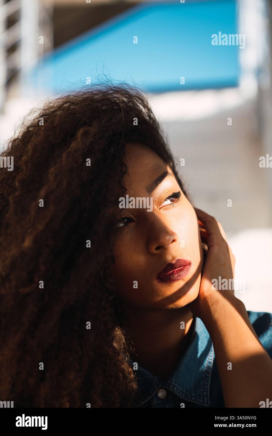 Ritratto di una donna nera premurosa che guarda lontano, scattata all'aperto con la luce naturale del sole. La composizione verticale e l'atmosfera luminosa lo rendono versatile per le campagne lifestyle e fashion. Ideale per storie di empowerment, stile quotidiano e vita positiva. Foto Stock