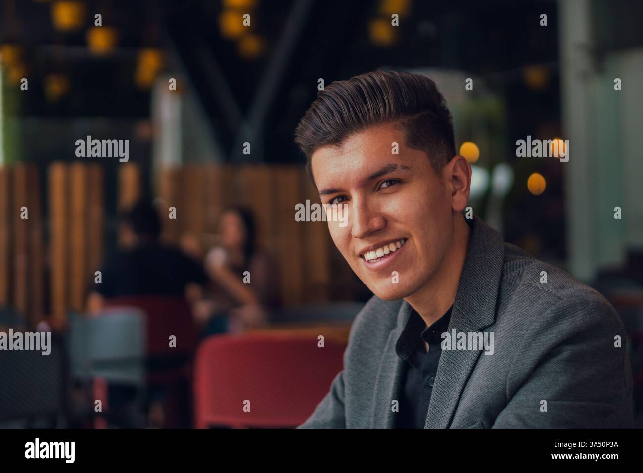 Ritratto di un giovane uomo positivo in una tuta elegante sorridente alla macchina fotografica mentre si siede in un bar moderno. Situata in una lounge contemporanea, questa immagine supporta contesti di lavoro, lifestyle e profili professionali. Ideale per comunicazioni aziendali, moda o ristoranti. Foto Stock