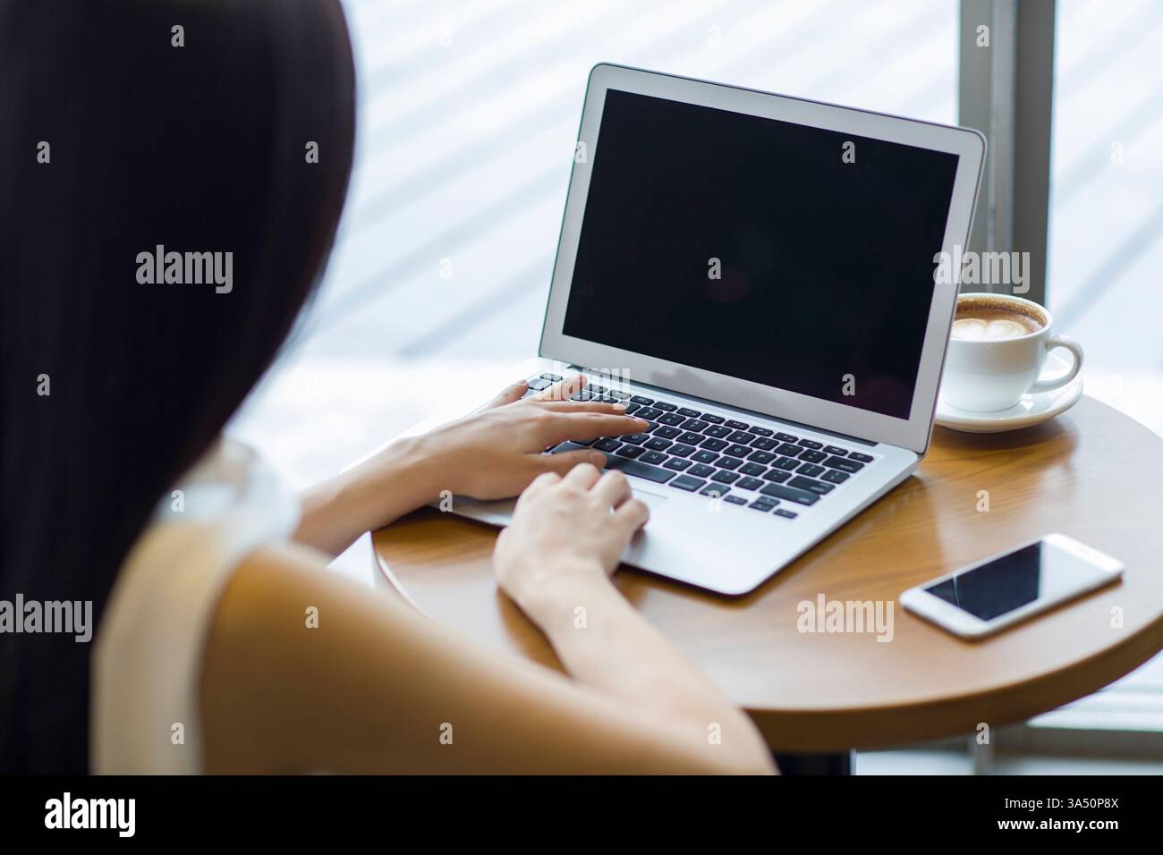 Donna d'affari cinese che lavora su un portatile in un bar, concentrata al tavolo. Ideale per storie di lavoro urbano, tecnologia e affari informali in un ambiente cittadino. Foto Stock