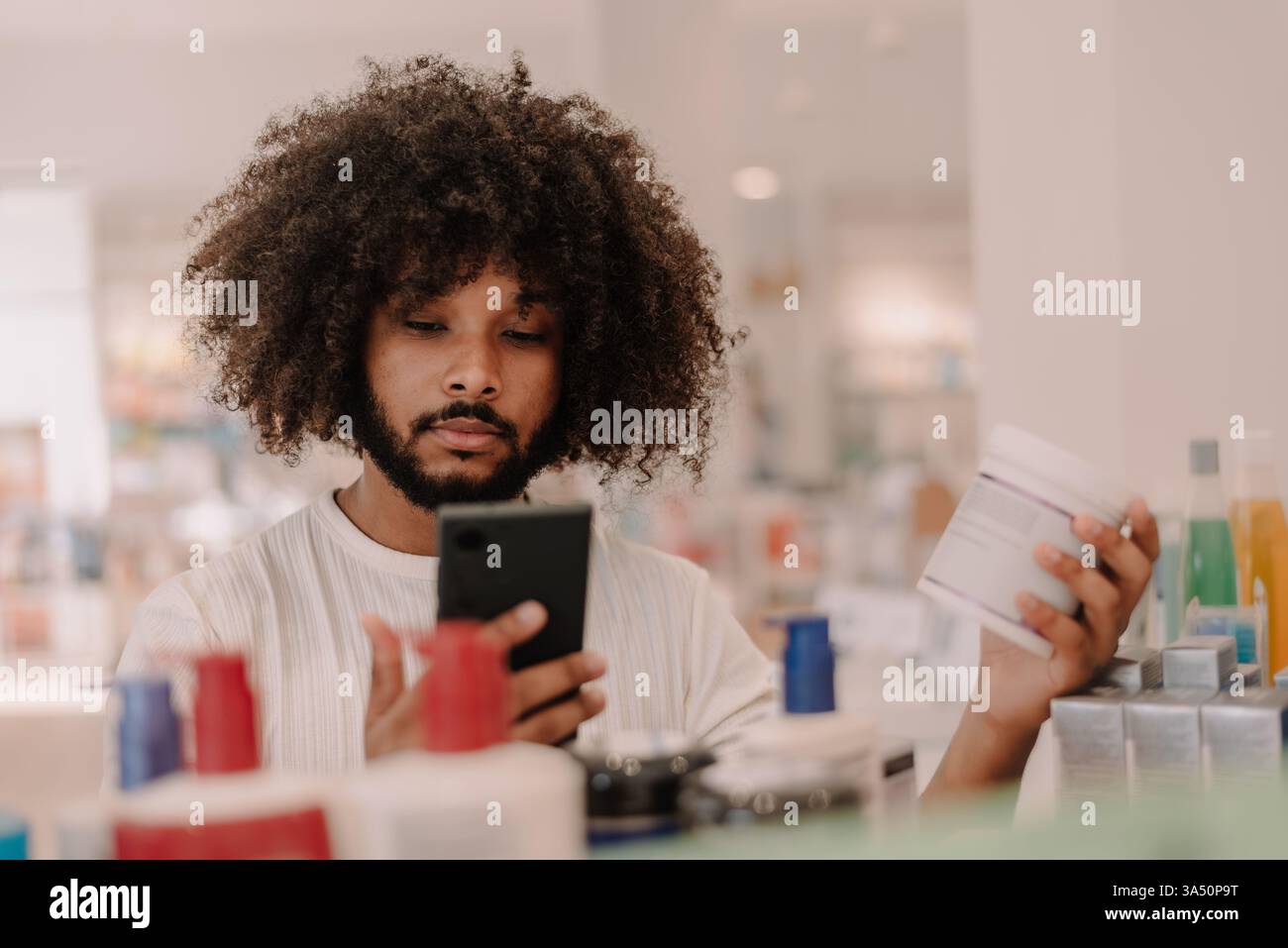 Uomo multirazziale con barba afro controlla le informazioni sui farmaci sul suo smartphone accanto agli scaffali della farmacia. Ideale per argomenti come la farmacia online, la guida ai farmaci e lo shopping sanitario. Foto Stock