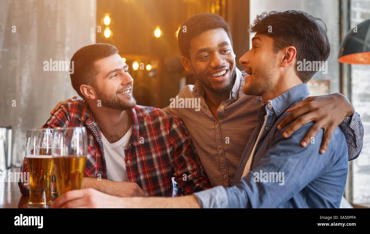 Un uomo nero sorridente e amici caucasici che parlano seduti insieme a tavola con un bicchiere di birra al bar Foto Stock
