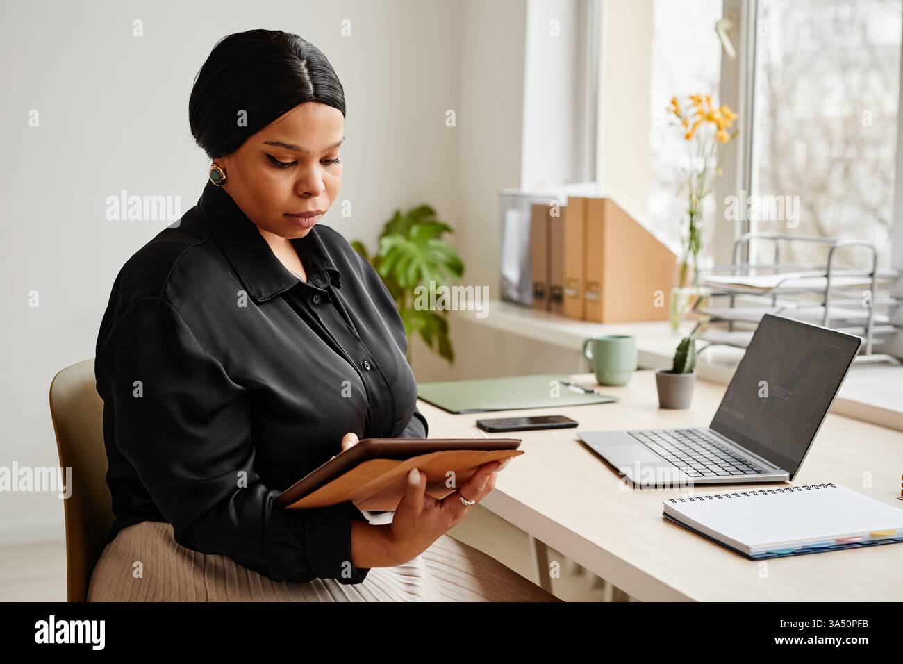 Ritratto di un'elegante donna d'affari nera che usa un tablet alla scrivania in un ambiente minimalista. L'ambiente pulito e moderno include spazio di copia per il branding. Perfetto per il branding aziendale, le donne nelle aziende e le campagne sul digital workplace. Foto Stock