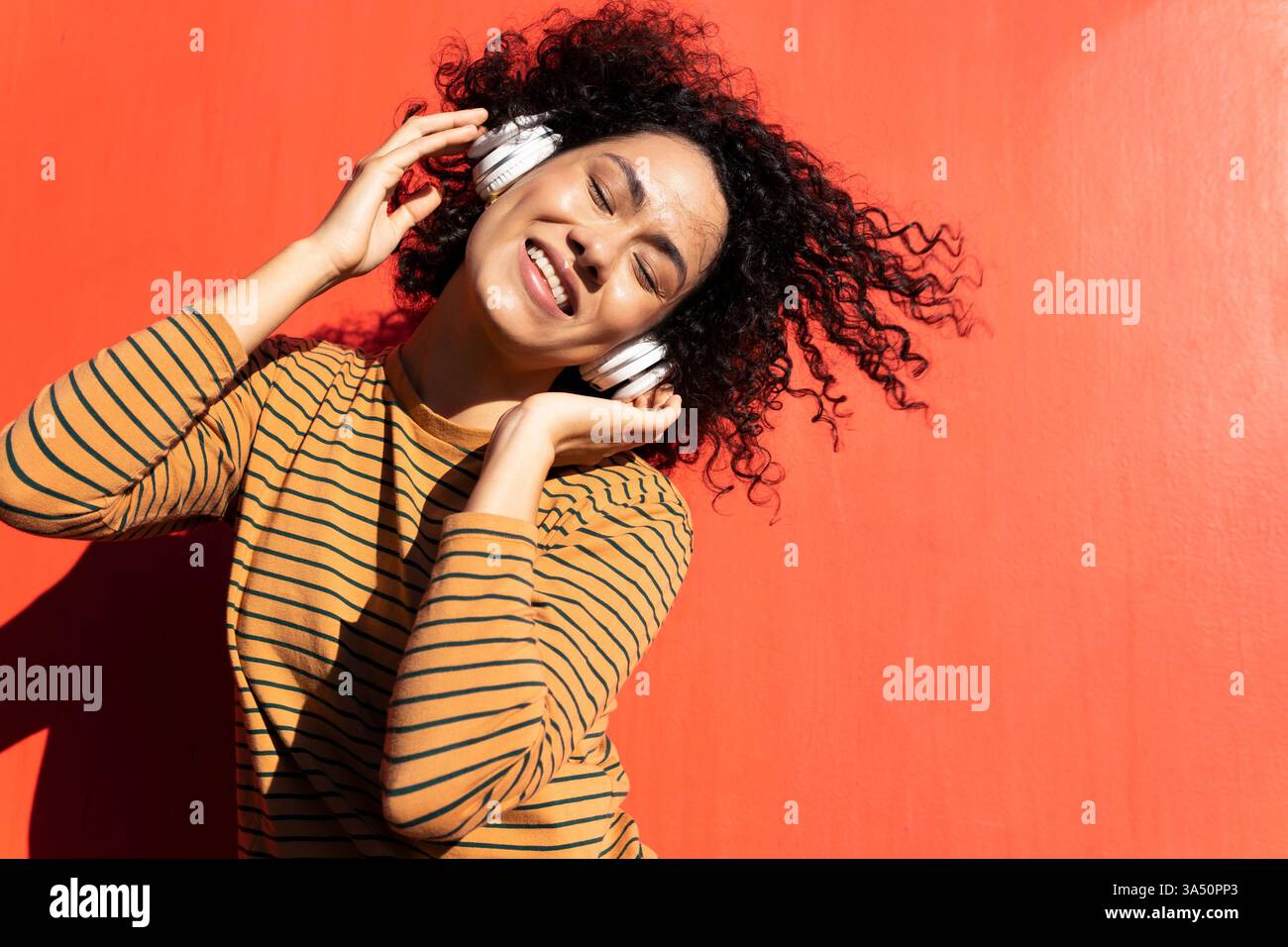 La donna ispanica che indossa le cuffie ama la musica con gli occhi chiusi. L'atmosfera luminosa e colorata si adatta a temi di stile di vita e intrattenimento. Un'immagine versatile per la musica, il relax e le campagne di benessere. Foto Stock