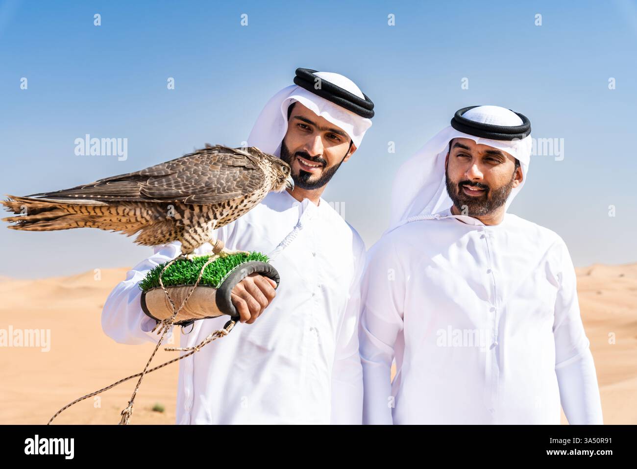 Due uomini mediorientali in tradizionali kandura legano nel deserto, tenendo un falco. Questo sorprendente momento culturale cattura l'amicizia, il patrimonio culturale e la falconeria in un ambiente desertico. Ideale per viaggi in Medio Oriente, cultura e stili di vita. Foto Stock