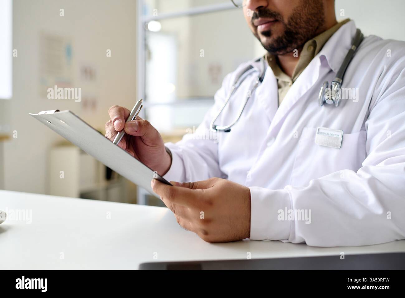 Primo piano di un medico con un cappotto bianco che scrive una prescrizione alla sua scrivania nell'ufficio della clinica. Il momento focalizzato mette in evidenza l'assistenza medica, la consultazione dei pazienti e la professionalità sanitaria. Ottimo per la pratica medica, farmacia, o informazioni sulla salute visuali. Foto Stock