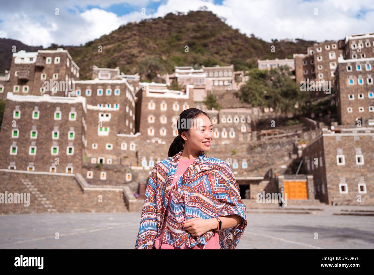 La donna turistica asiatica si trova davanti agli edifici storici in pietra del villaggio di Rijal Alma, in Arabia Saudita. Questa immagine della destinazione mette in risalto la cultura, l'architettura e l'esplorazione del viaggio. Ottimo per il marketing turistico e le guide turistiche. Foto Stock