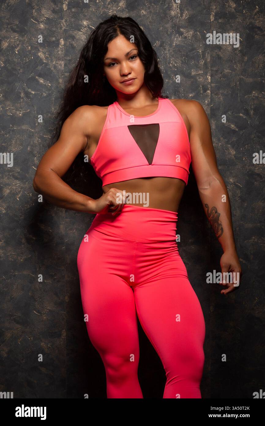 La culturista ispanica posa con la mano sull'anca contro una parete grigia. Il forte fisico e la posizione atletica mettono in risalto il fitness e il bodybuilding. Perfetto per fitness, sport e temi di empowerment. Foto Stock