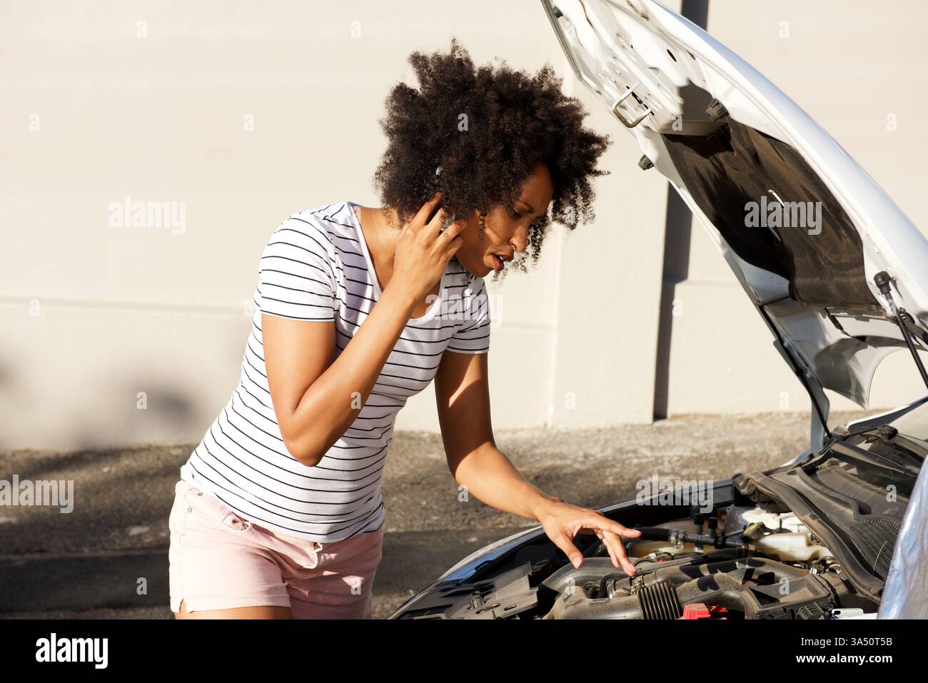 Ritratto di una giovane donna africana in piedi accanto a un'auto in avaria sulla strada, che chiede assistenza. La scena stradale all'aperto con uno smartphone in mano mette in risalto i problemi automobilistici e la ricerca di aiuto. Adatto per riparazioni auto, assistenza stradale e contesti di servizi di emergenza. Foto Stock