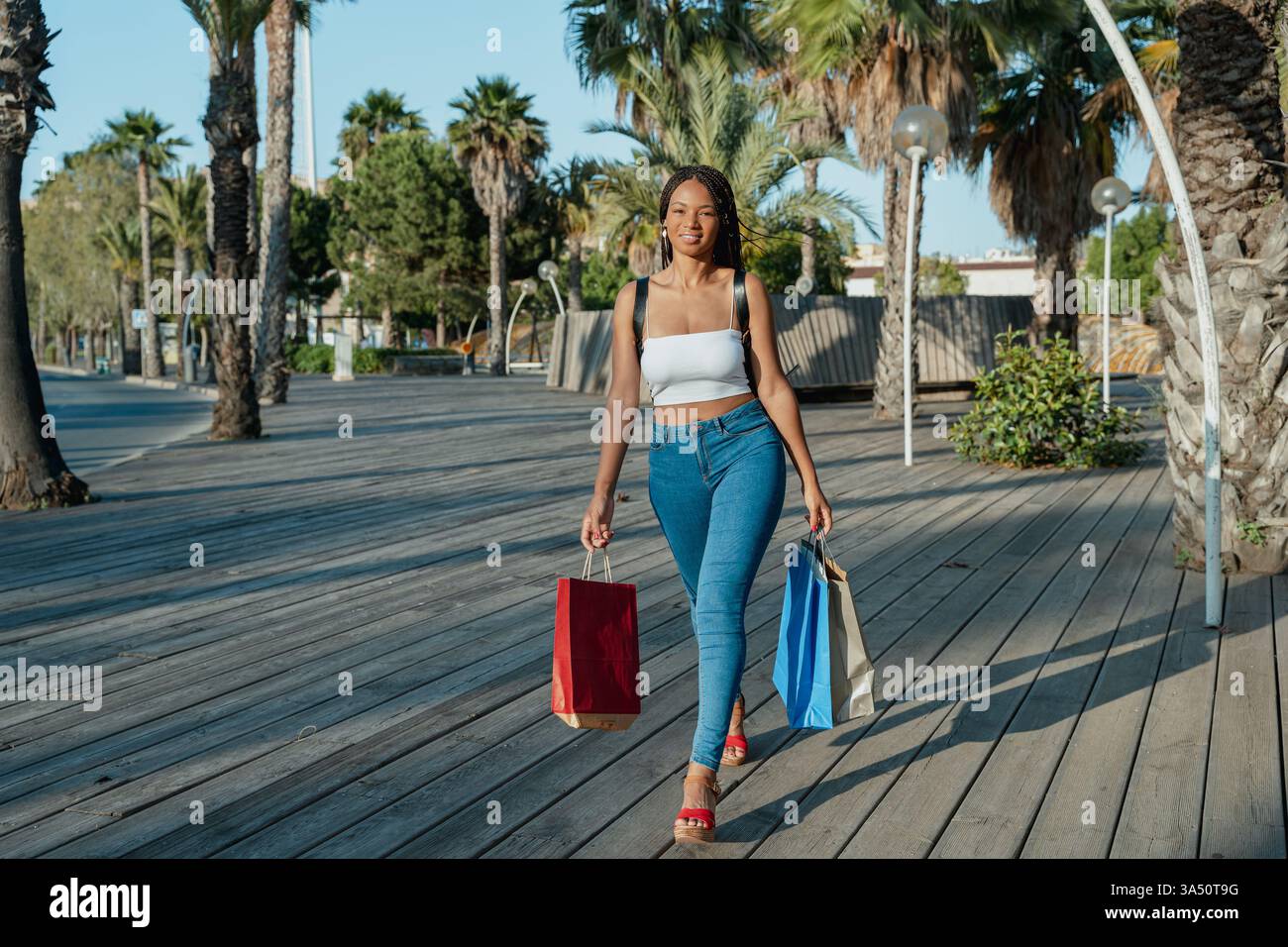 Un'elegante donna nera con borse per lo shopping cammina lungo una strada soleggiata della città. Questa vivace scena urbana è perfetta per lo stile di vita, la moda e i momenti di consumo. Foto Stock