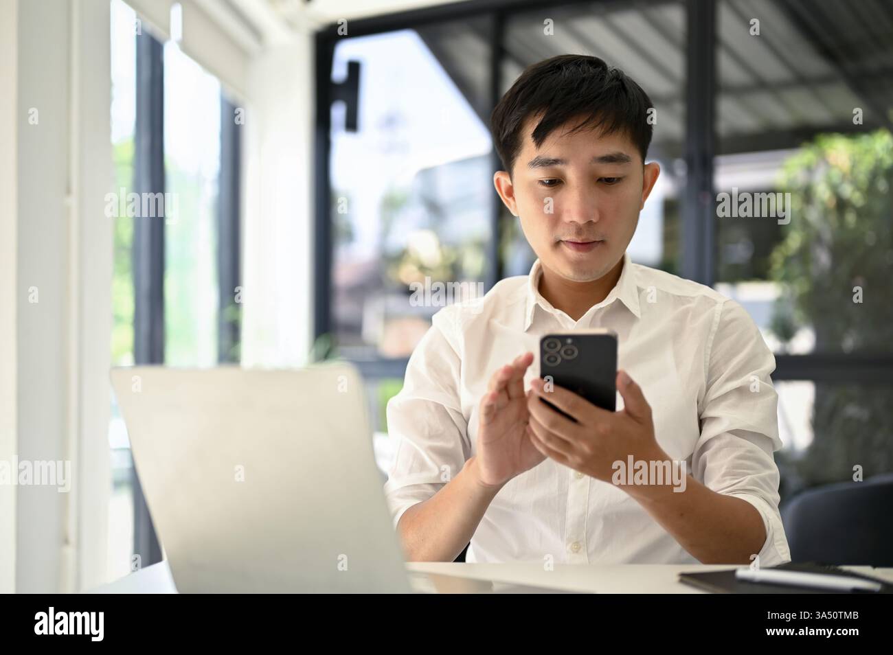 L'uomo d'affari asiatico millenario controlla i messaggi sul suo smartphone alla sua scrivania in ufficio. Ritratto aziendale contemporaneo incentrato su tecnologia, comunicazione e produttività. Foto Stock