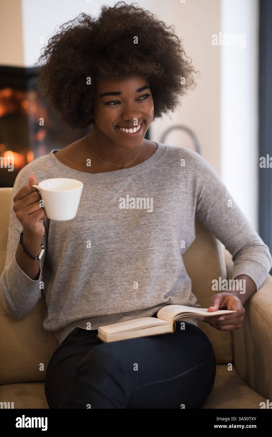 Allegra donna nera con un afro seduto su un divano del soggiorno, tenendo in mano una tazza di caffè e un libro accanto al caminetto. Il caldo ambiente interno offre un confortevole relax a casa e un'atmosfera autunnale. Foto Stock