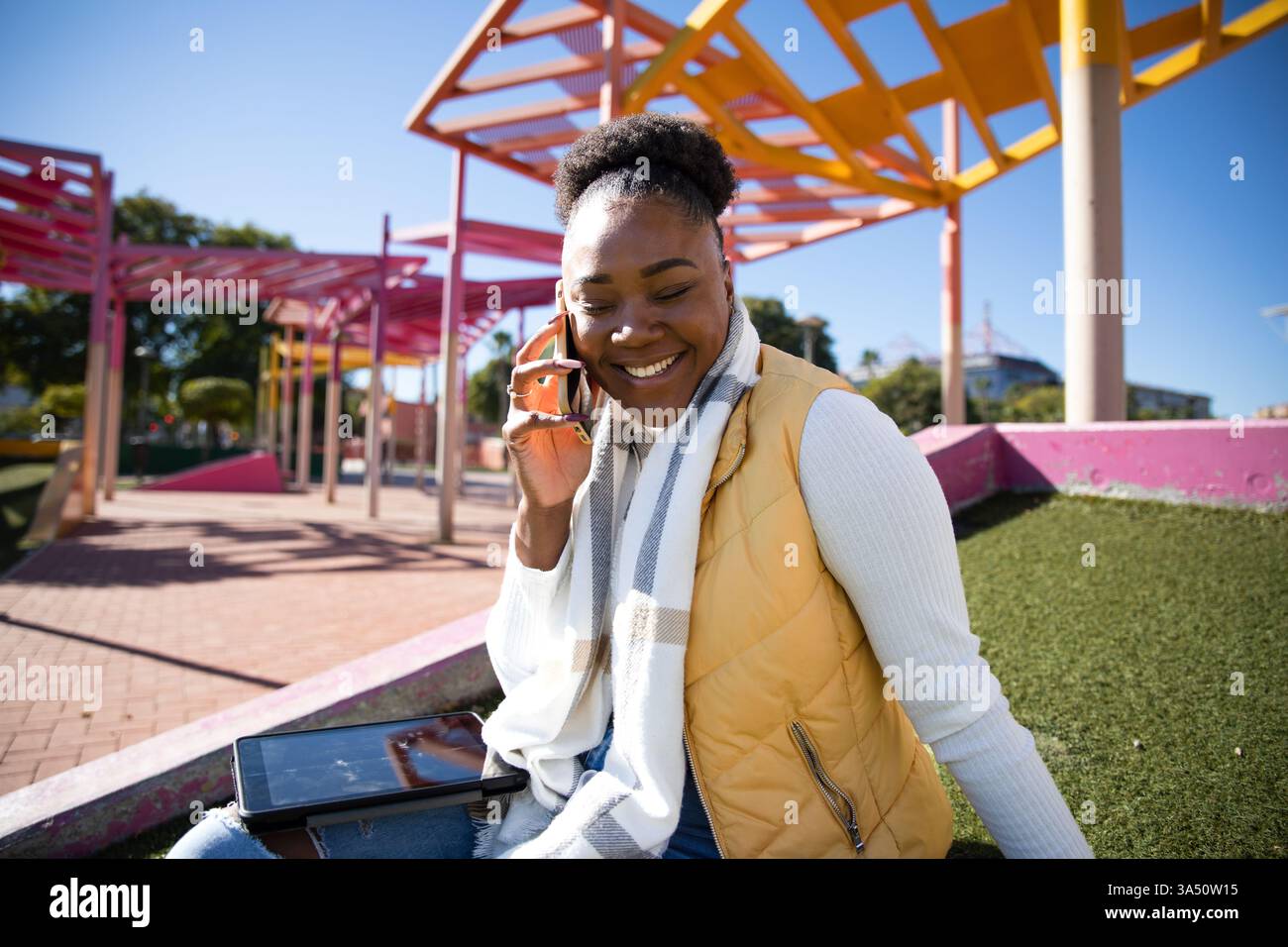 Una allegra donna afroamericana siede in un parco cittadino in una giornata di sole, parlando al telefono. Possiede un tablet, evidenziando uno stile di vita connesso e mobile. Ideale per la vita urbana, la comunicazione digitale e i temi del lavoro da remoto. Foto Stock