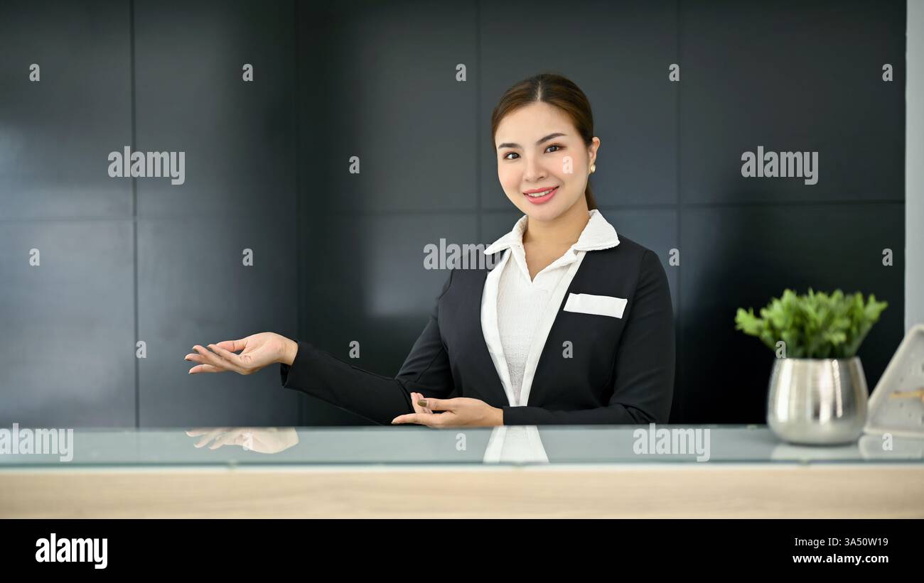 Accogliente e attraente receptionist femminile asiatica millenaria in un'uniforme si erge presso il banco del ricevimento nella lobby moderna. Concetto di servizio di ospitalità Foto Stock