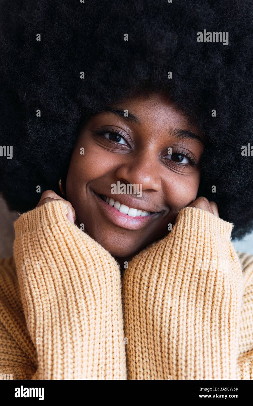 Ritratto di una allegra donna afroamericana con acconciatura afro su sfondo grigio. Le sue mani sono infilate all'interno di un comodo maglione mentre riposa il viso, irradiando calore e calma. Foto Stock