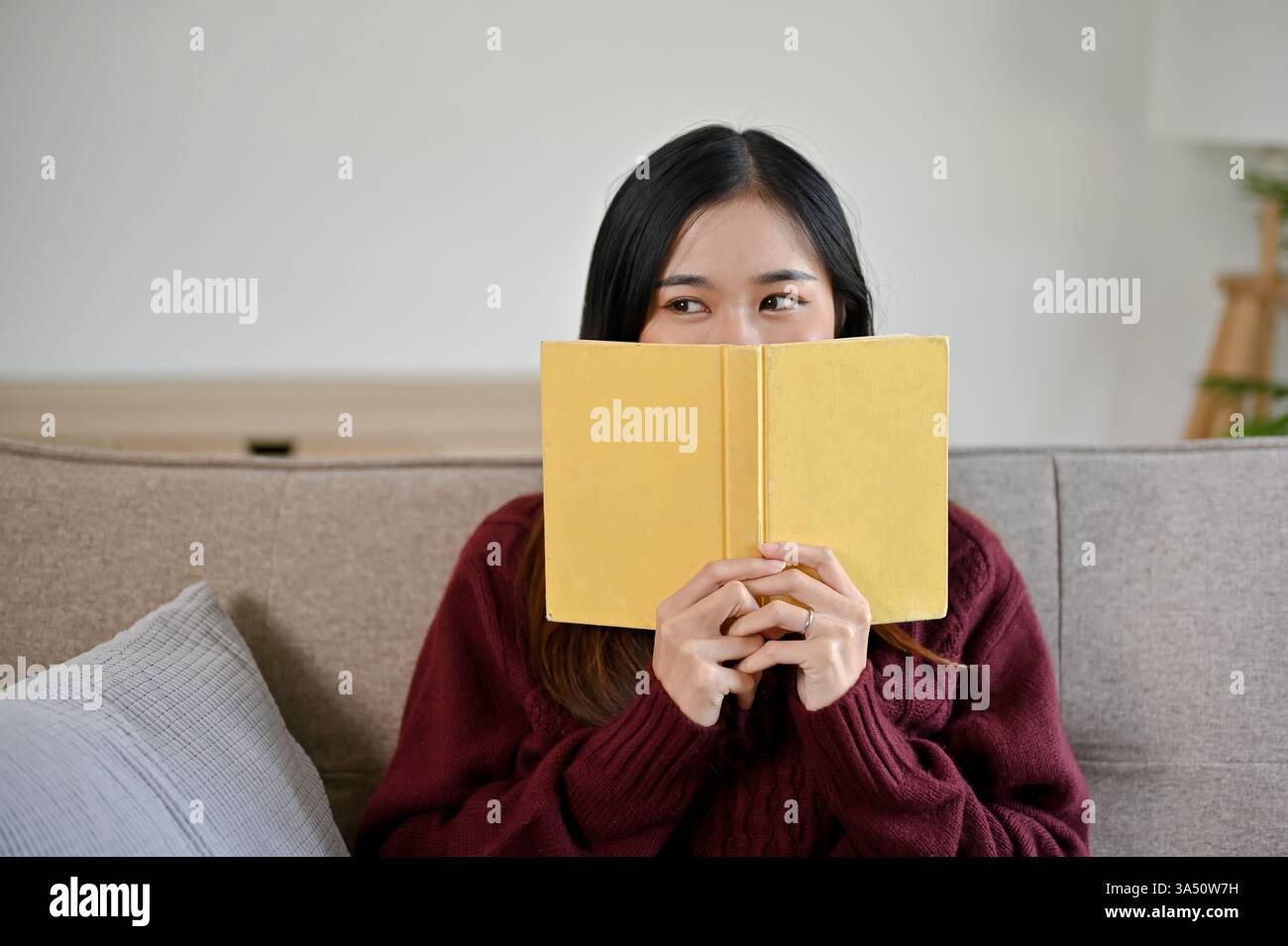 Donna asiatica in maglione rosso siede su un divano, coprendo il suo viso con un libro mentre si rilassa. Un momento accogliente, ideale per leggere, rilassarsi o dedicarsi a contenuti di stile di vita. Perfetto per scene comode da salotto e campagne di autoassistenza. Foto Stock