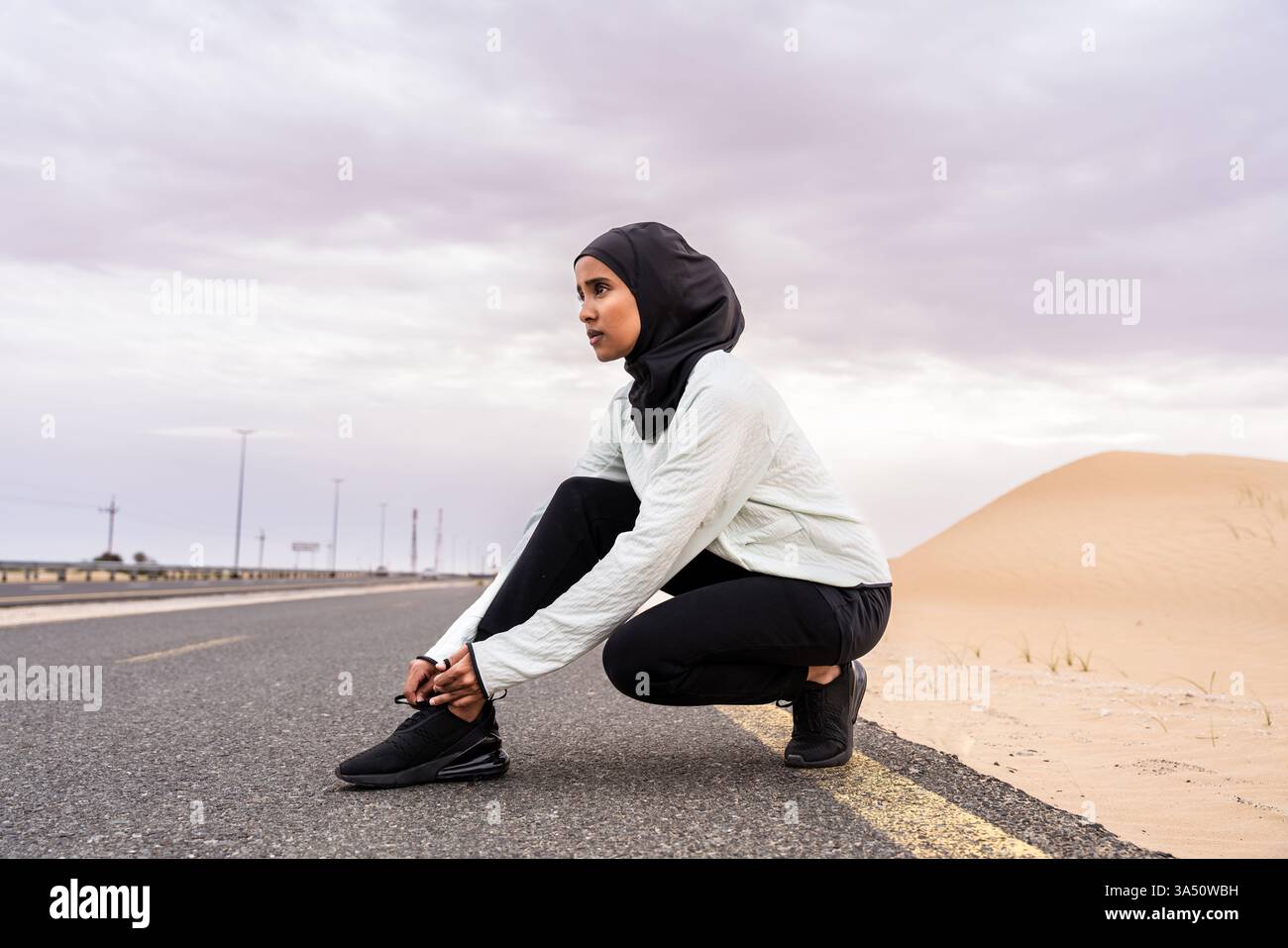 Donna atletica mediorientale che indossa un hijab all'aperto in un ambiente desertico. Questo ritratto di stile atletico mette in evidenza fitness, determinazione e attività fisica all'aperto in un deserto soleggiato. Foto Stock