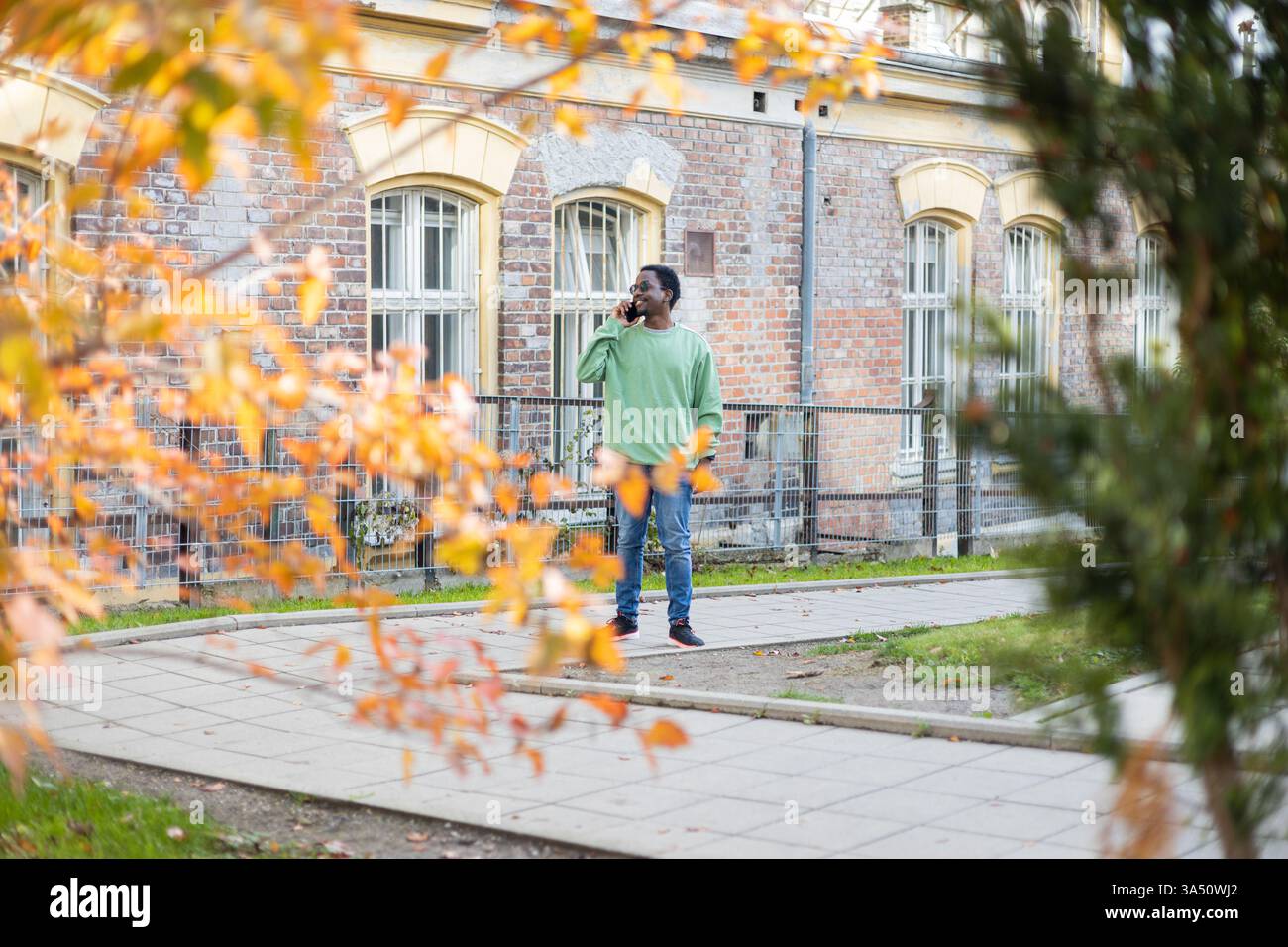 Vista laterale di un uomo afroamericano sorridente che parla su un telefono cellulare mentre si trova vicino a un edificio su un passaggio pedonale pavimentato. L'ambiente urbano con luce diurna trasmette uno stile di vita attivo e professionale. Ideale per temi aziendali, tecnologici o pendolari. Foto Stock