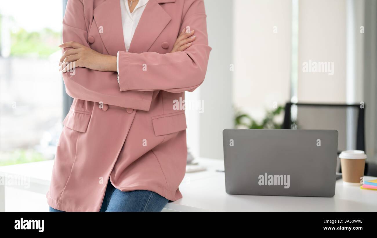 Donna d'affari sicura con un abito rosa siede a una scrivania con le braccia incrociate, presentando un'immagine aziendale avvolta. Ideale per le funzioni di donne in affari, leadership e presenza dirigenziale. I ritratti ritagliati sono ideali per le intestazioni e gli spazi di copia nei materiali di marketing professionali. Foto Stock