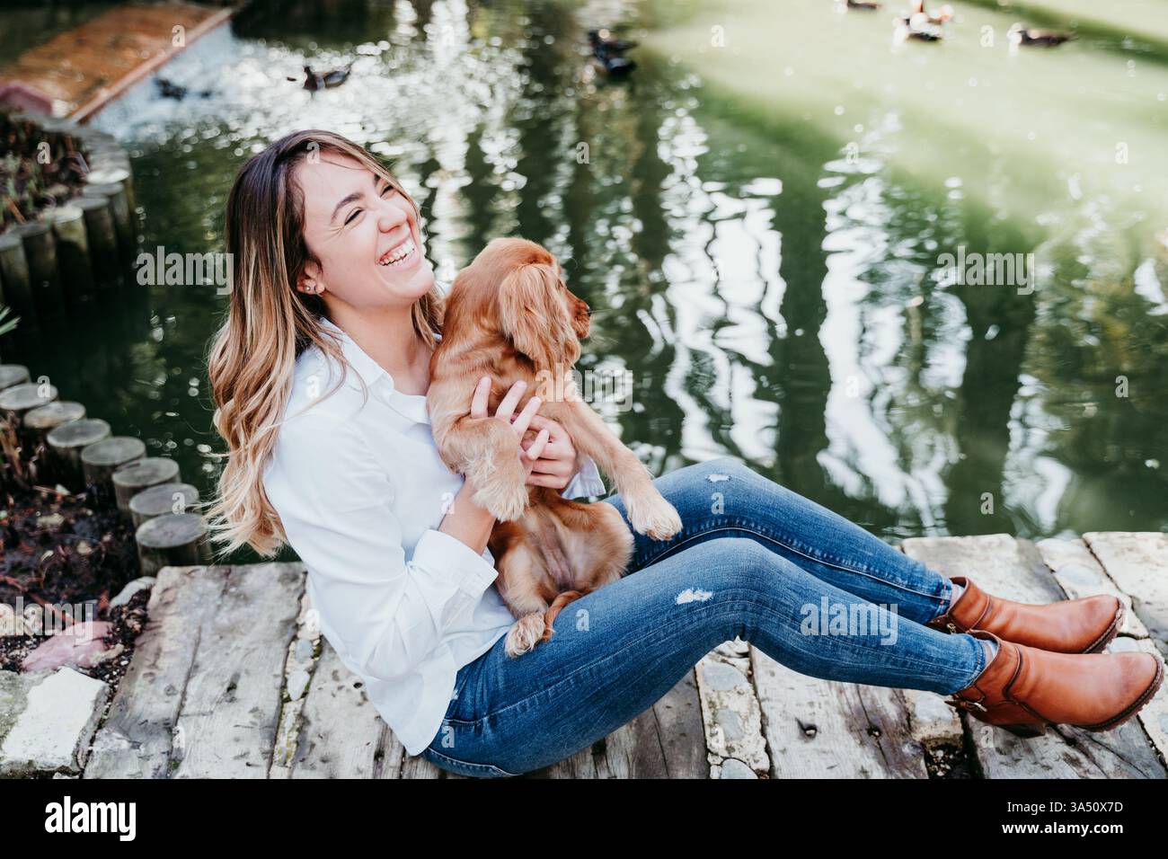 Allegra donna ispanica siede con il suo cane su un passaggio pedonale in legno vicino a uno stagno durante il giorno. Questa immagine di stile di vita all'aperto per animali domestici si adatta alle campagne in cui sono ammessi i cani. Foto Stock