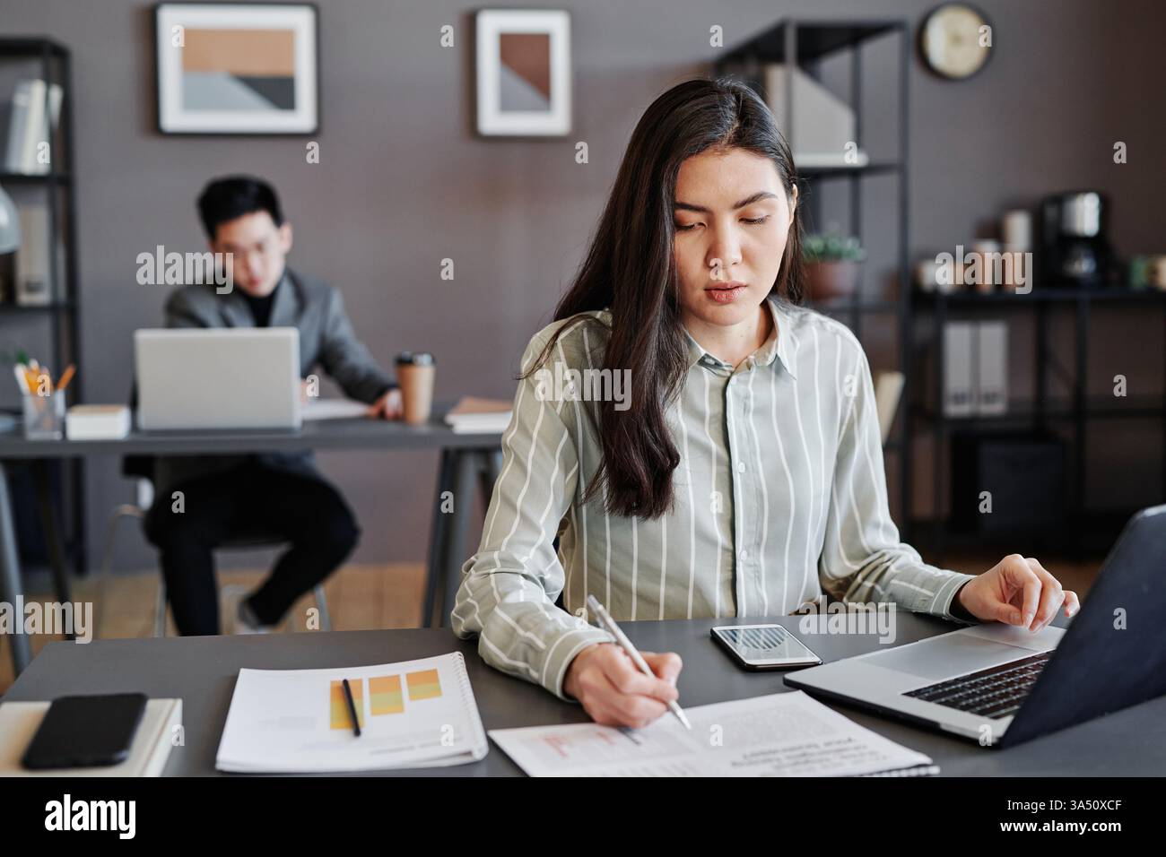 Immagine orizzontale di una giovane donna asiatica a una scrivania in un ufficio, che lavora con grafici e grafici. Una foto lucida per la finanza, l'analisi dei dati e le presentazioni aziendali. Adatto per relazioni con gli investitori, ricerche di mercato e comunicazioni aziendali. Foto Stock