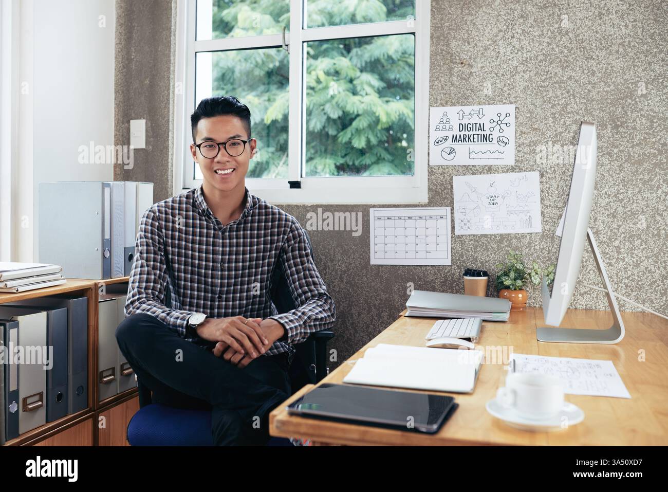 Il giovane imprenditore sorridente in occhiali si siede su una scrivania moderna con calendario e idee di avvio sul muro. Ideale per aziende, startup, tecnologia e stili di vita professionali. Foto Stock