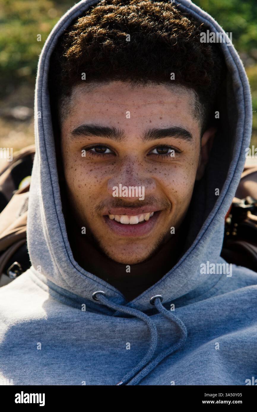 Un giovane ispanico sorridente dai capelli scuri giace su una strada urbana con una felpa con cappuccio grigia, che guarda la fotocamera con un look serio e premuroso. L'ambiente cittadino aggiunge stile di strada e atmosfera giovanile contemporanea. Adatto per la fotografia di strada, lo stile di vita urbano e i ritratti informali. Foto Stock