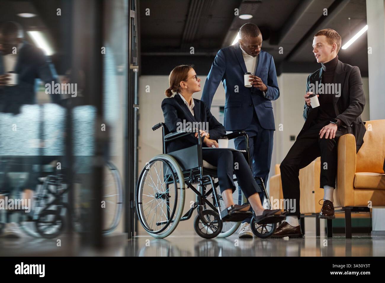 Ritratto a figura intera di un team aziendale diversificato in un ufficio moderno, con una giovane donna in sedia a rotelle che chiacchiera con colleghi maschi. Un ambiente di lavoro inclusivo che mette in evidenza l'accessibilità e il lavoro di squadra. Foto Stock