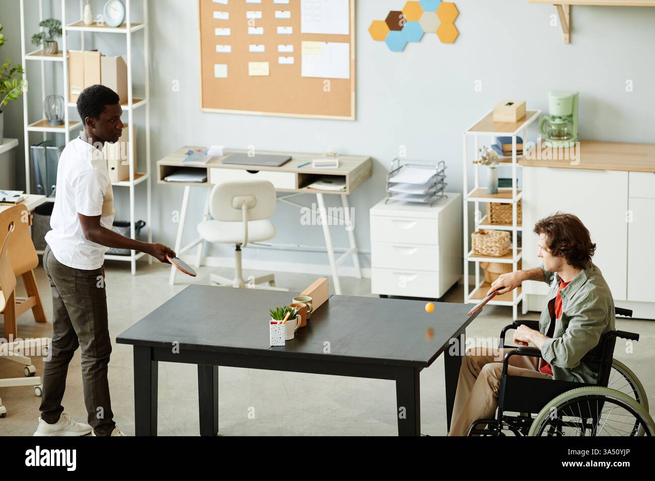 Vista laterale ritratto di un giovane con disabilità che gioca a ping-pong in una sala ristoro. La scena trasmette una cultura inclusiva sul luogo di lavoro e attività ricreative occasionali, con spazio di testo per il branding. Foto Stock