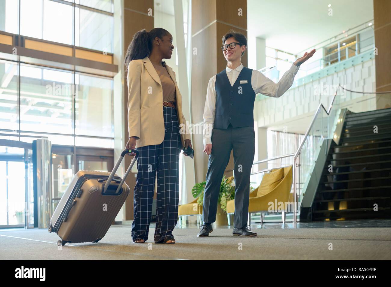 Un receptionist asiatico di sesso maschile che indossa occhiali che mostrano la strada per una donna d'affari di colore che cammina con i bagagli nella lobby dell'hotel Foto Stock