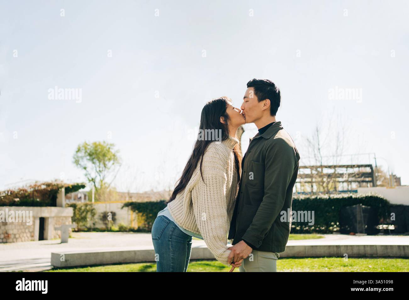 Coppia asiatica che si tiene per mano e si bacia in un parco cittadino, gli occhi chiusi e sorridenti. Un momento romantico in un ambiente urbano all'aperto. Adatto per campagne di amore, relazioni e stile di vita. Foto Stock