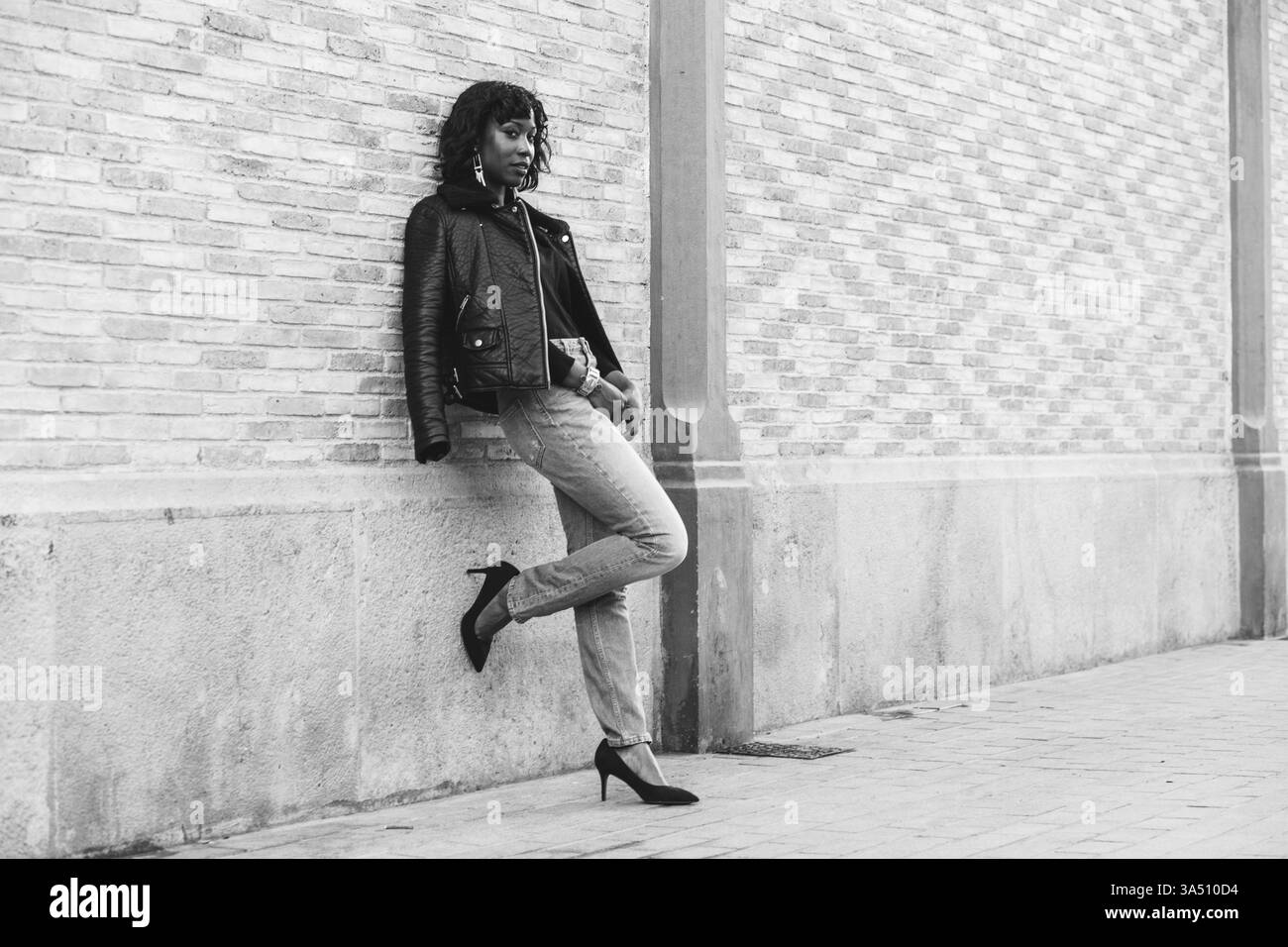 Donna afroamericana appoggiata contro un muro di mattoni su una strada cittadina. L'immagine trasmette stile di strada, sicurezza e moda urbana. Foto Stock