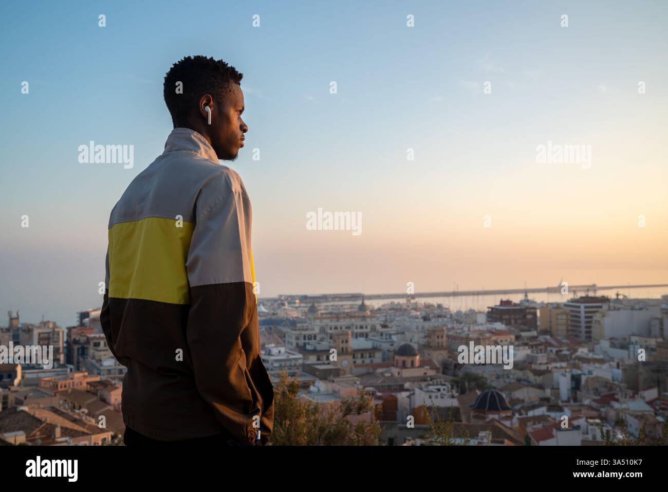 Vista laterale di un giovane uomo sicuro di sé che indossa cuffie senza fili, ammirando una pittoresca città costiera contro un cielo senza nuvole. Perfetto per stili di vita, viaggi e musica in contesti urbani moderni. Atmosfera pulita e rilassata con particolare attenzione alla tecnologia wireless. Foto Stock