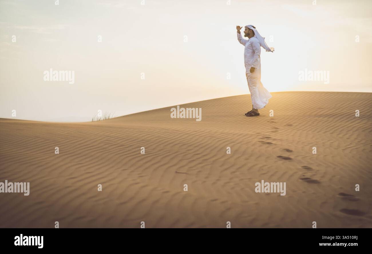 Un serio uomo mediorientale in abiti tradizionali degli Emirati si trova in un deserto all'alba, guardando in lontananza. Un'immagine sorprendente per i viaggi, la cultura e i paesaggi desertici del Medio Oriente. Adatto per temi di esplorazione, solitudine e viaggio in ambienti aridi. Foto Stock