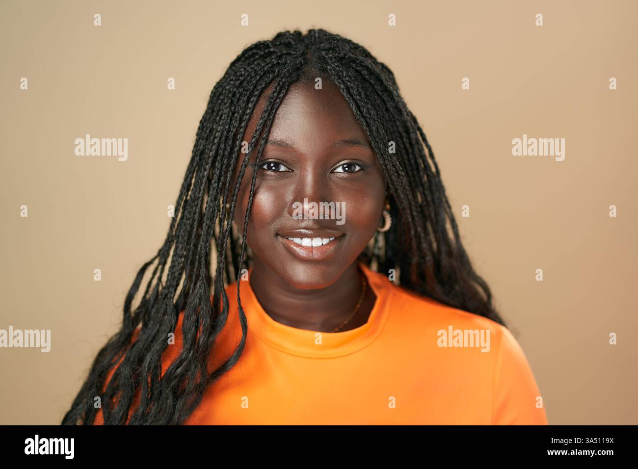 Ritratto di una donna afroamericana sorridente con trecce afro che indossa un top arancione, in piedi su uno sfondo marrone da studio. Il suo sguardo sicuro e i colori vivaci rendono questa headshot ideale per campagne di moda, bellezza e diversità. Foto Stock