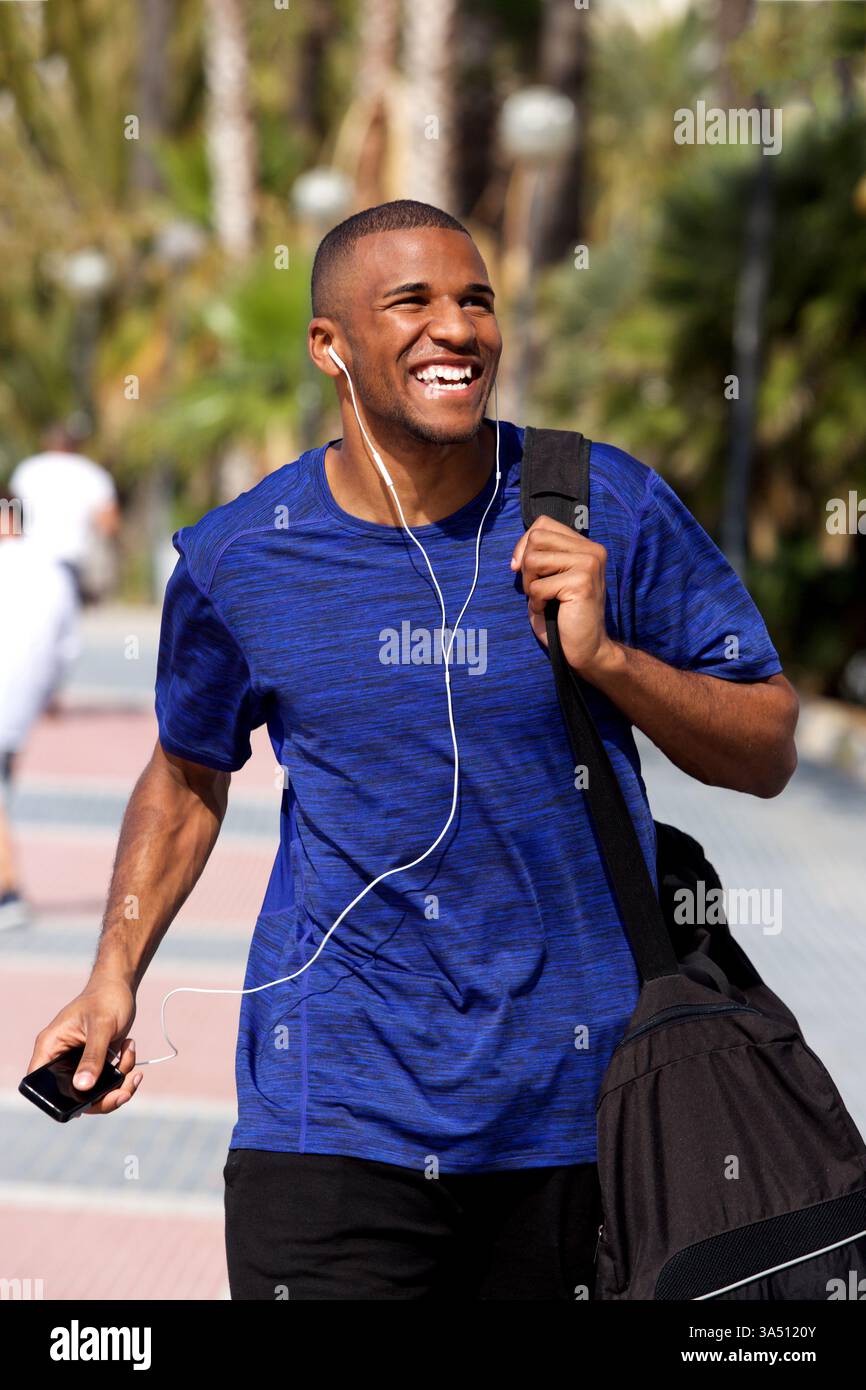 Allegro giovane sportivo africano che cammina all'aperto con una borsa sportiva. L'immagine trasuda vitalità e uno stile di vita attivo in un ambiente urbano o in un parco. Ideale per il branding di sport, fitness e streetwear. Foto Stock