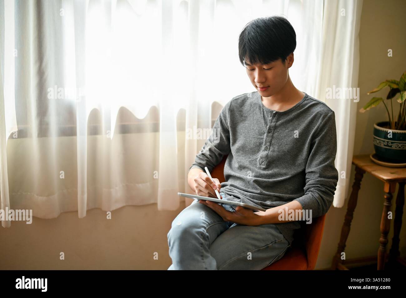 Uomo asiatico che usa uno stilo su un tablet in un ambiente domestico informale. Si siede su una sedia con le gambe incrociate accanto a una finestra, catturando un momento di vita al chiuso diurno. Questa immagine è adatta ad argomenti come il disegno digitale, l'apprendimento online e lo stile di vita tecnologico. Foto Stock