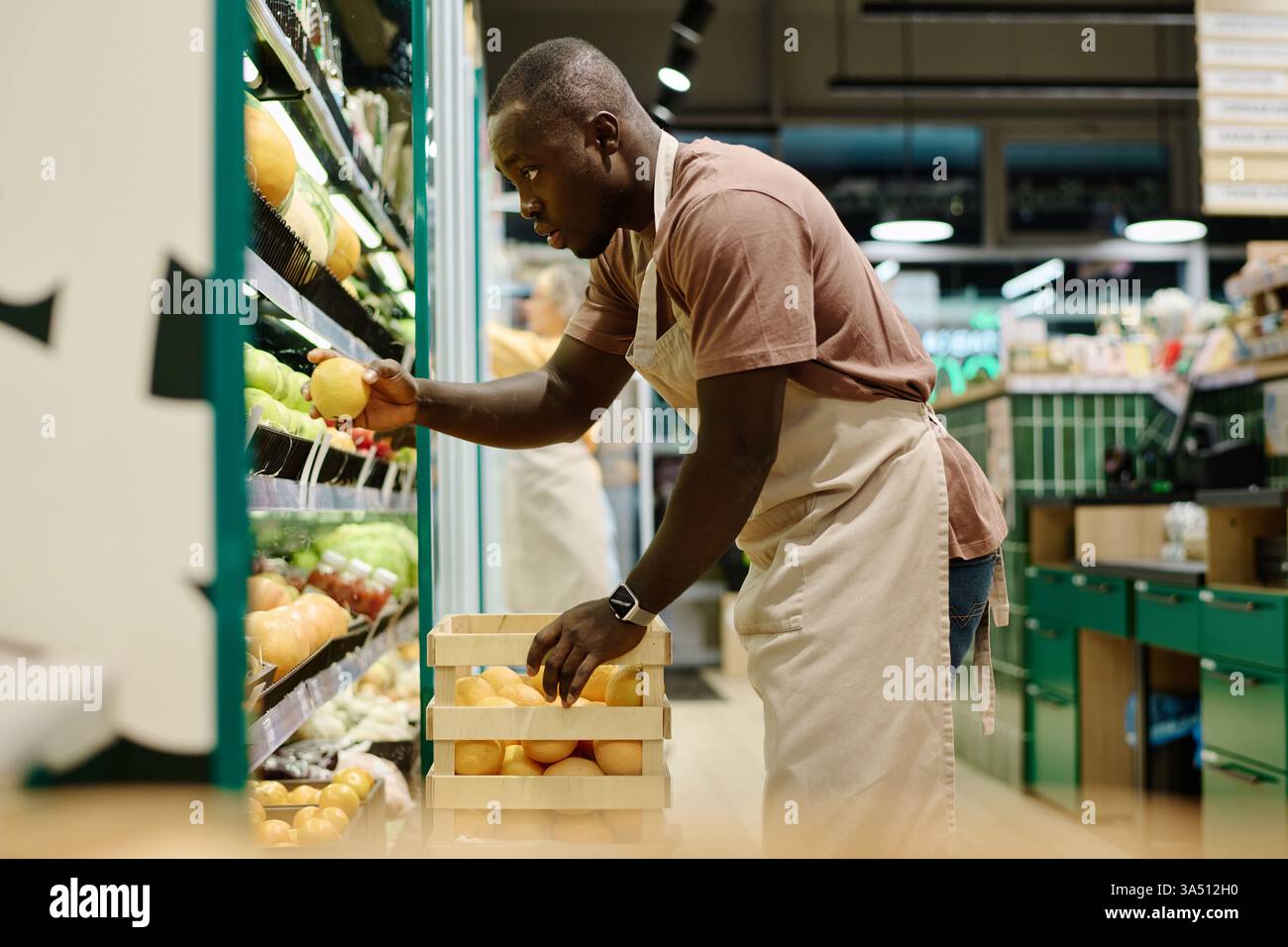 Mercante afroamericano che organizza frutta fresca sugli scaffali dei supermercati. Ideale per la vendita al dettaglio, la drogheria e le immagini di negozi alimentari. Foto Stock