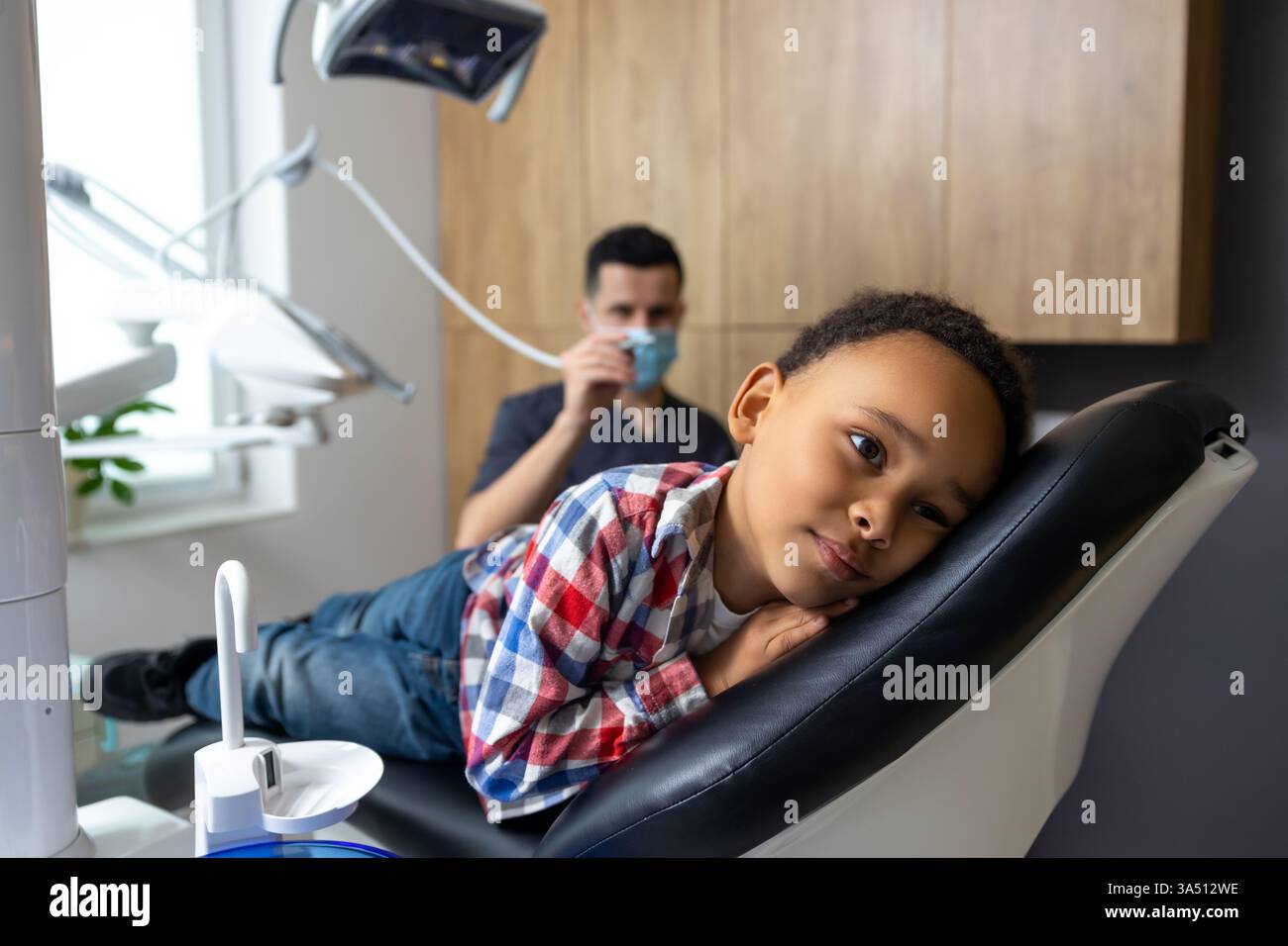 Bambino dalla pelle scura sdraiato su una sedia dentale all'interno dell'ufficio di un dentista durante un check-up di routine. La scena mette in evidenza un ambiente odontoiatrico pediatrico dotato di apparecchiature odontoiatriche e lampada clinica. Foto Stock