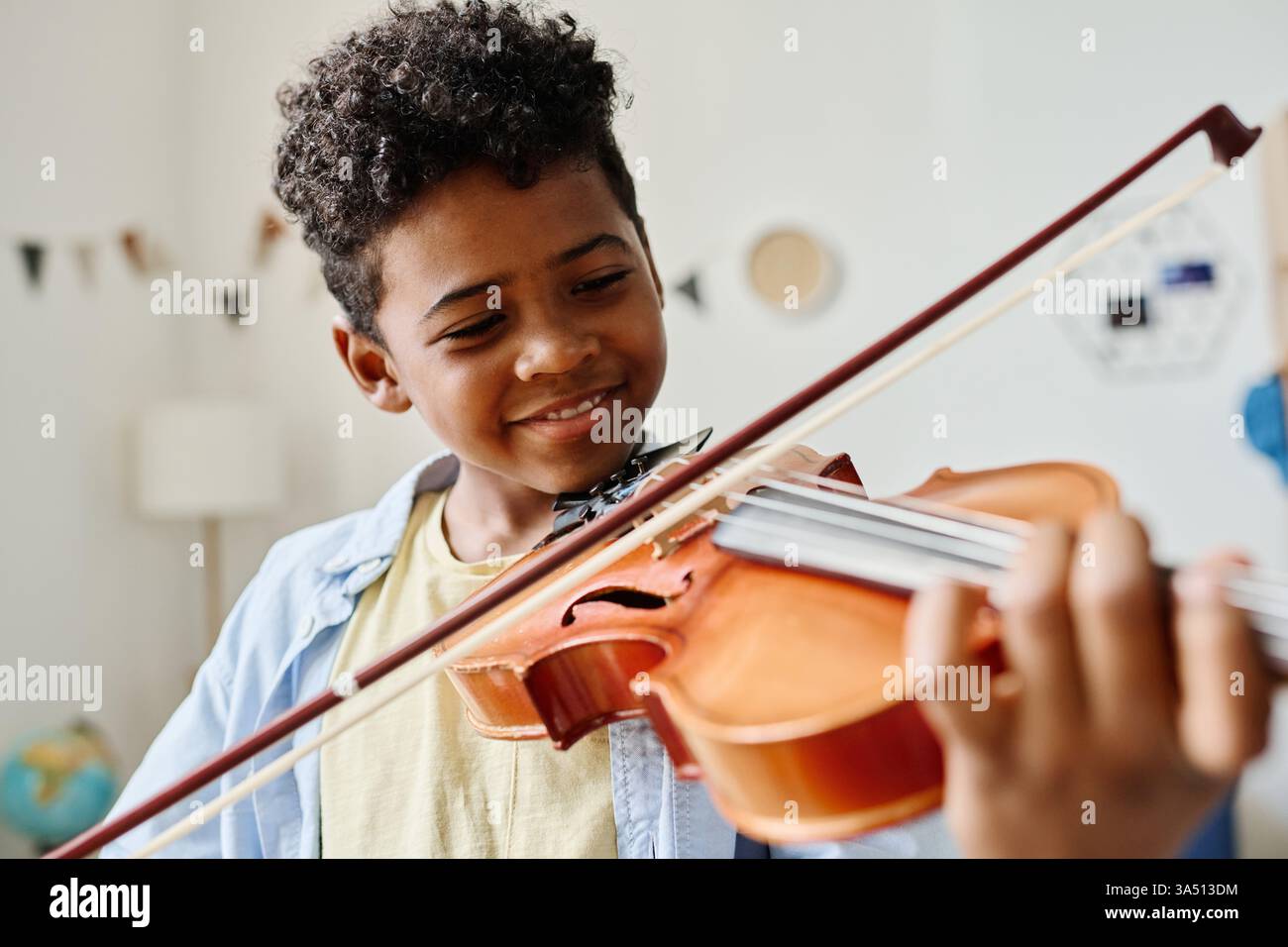 Sorridente ragazzo afroamericano suona il violino a casa durante una lezione di musica. Ottimo per contenuti sull'educazione musicale dei bambini, sull'apprendimento del violino e sulla pratica domestica. Cattura un momento caldo di studio in un ambiente domestico. Foto Stock