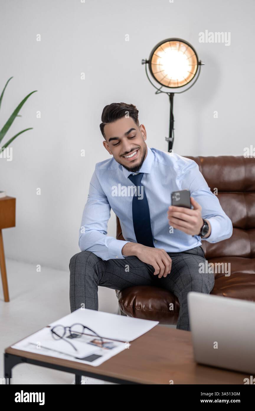 Il giovane uomo d'affari sorride mentre guarda il suo smartphone in un ufficio moderno. Si trova in uno spazio di lavoro elegante, trasmettendo successo ed energia positiva. Questa foto è ideale per temi di comunicazione aziendale, di leadership e sul luogo di lavoro. Foto Stock