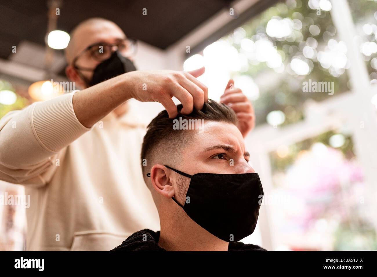 Barbiere professionale acconcia i capelli di un cliente all'interno di un barbiere pulito e moderno. La scena luminosa e mirata mette in risalto la cura del corpo e il taglio di precisione. Ideale per la cura del corpo maschile, la pubblicità nel barbiere e le immagini del salone post-pandemia. Foto Stock