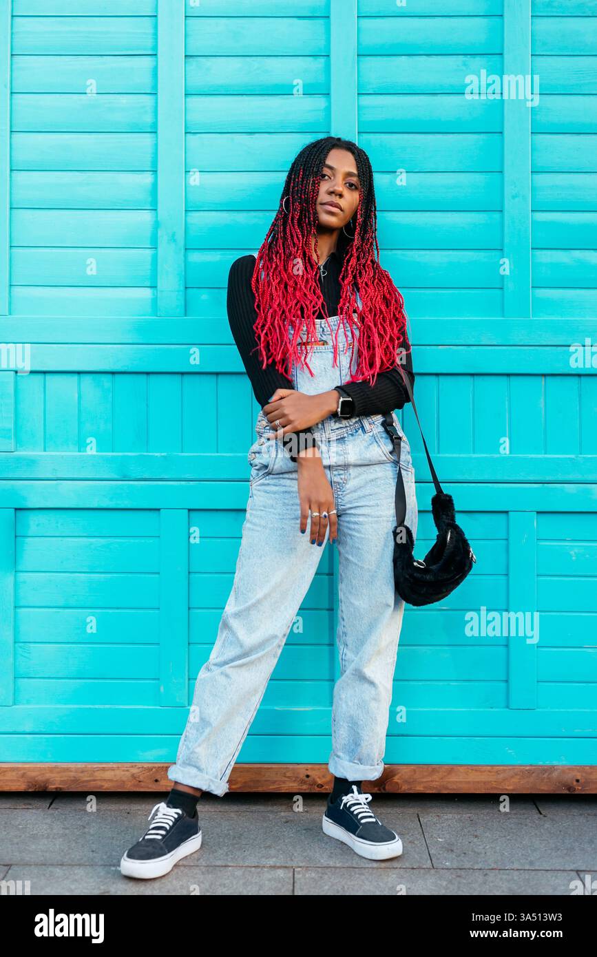 Donna nera sicura di sé in piedi all'aperto contro un muro blu. Questo ritratto Street style è perfetto per i temi della moda, dello stile di vita e dell'empowerment. Foto Stock