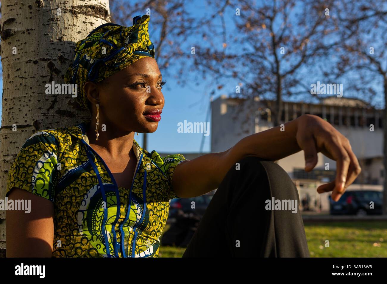 Donna nera che indossa una camicetta tradizionale e un velo siede in un parco cittadino soleggiato, guardando lontano. Questo autentico ritratto culturale cattura la vita quotidiana all'aperto con un cielo luminoso e un ambiente naturale. Ideale per collezioni fotografiche di stile di vita, diversità e cultura. Foto Stock