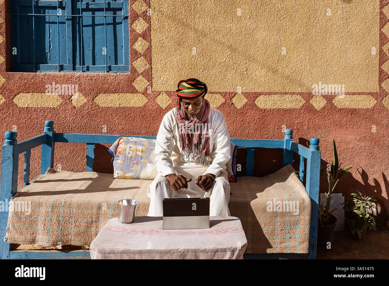 Sincero uomo egiziano in chat video tradizionali con turban e accappatoio su un tablet seduto su un divano in un cortile soleggiato. Una scena calda che fonde cultura e tecnologia, adatta per i momenti in famiglia e la connessione online. Ideale per storie di stile di vita sul lavoro a distanza, la vita a casa e il patrimonio culturale. Foto Stock
