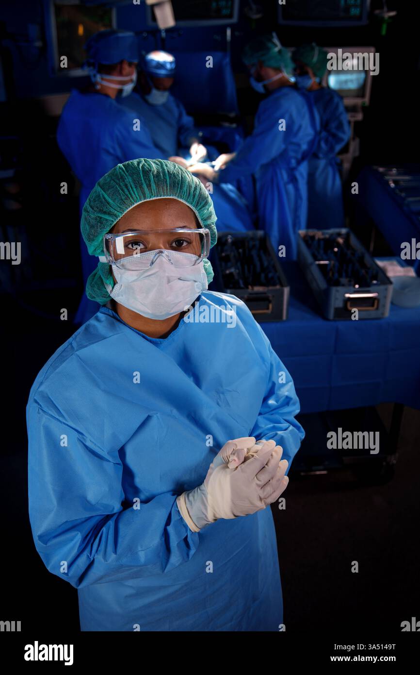 Ritratto di un'infermiera afroamericana in prima linea in scrub all'interno di una sala operatoria dell'ospedale con un team medico. Ideale per campagne di assistenza sanitaria, infermieristica e assistenza ospedaliera incentrate su operatori in prima linea e lavoro di squadra clinico. Ideale per rapporti e articoli su professionisti medici e cura dei pazienti. Foto Stock
