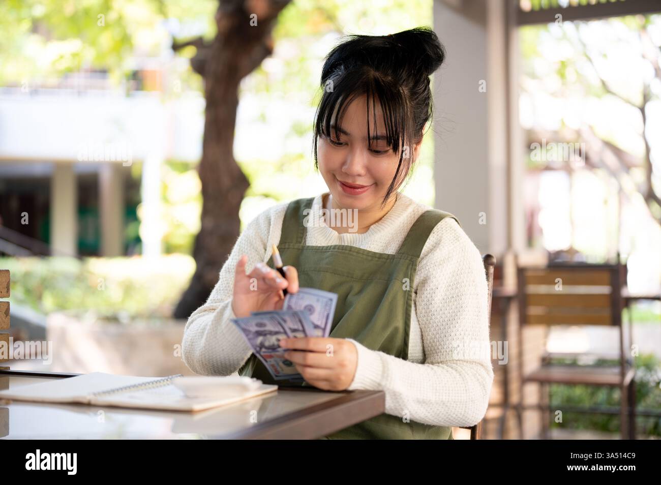 Felice giovane donna asiatica proprietaria di piccole imprese che conta i soldi e rivede le sue entrate mensili. Questa autentica scena imprenditoriale mette in evidenza la gestione del contante, il budget e la proprietà del negozio. Ideale per argomenti relativi all'imprenditoria femminile, alla finanza per le piccole imprese e alla crescita dei ricavi. Foto Stock