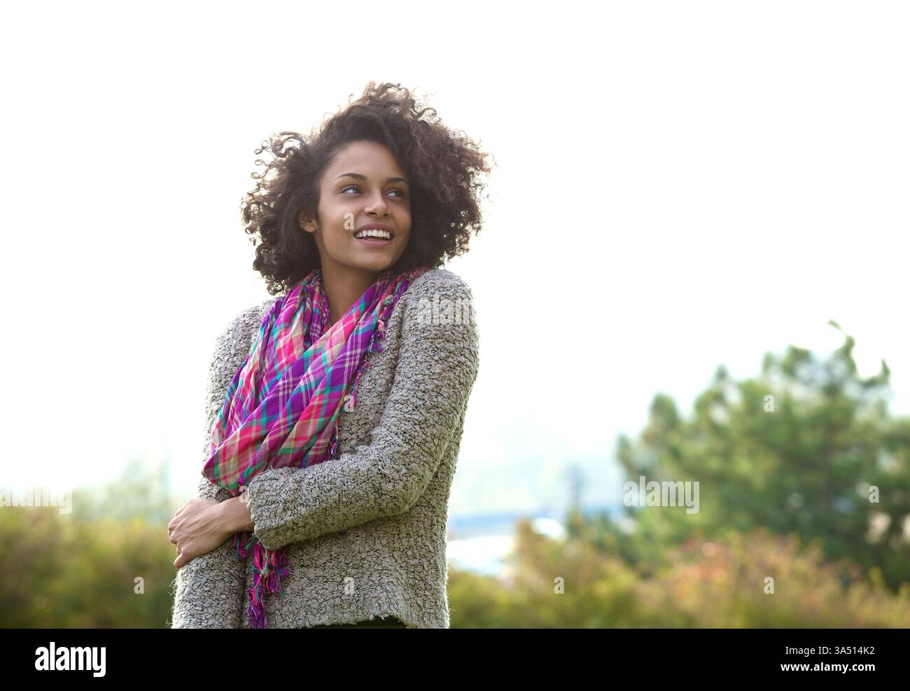 Donna afroamericana sorridente con afro in piedi vicino a piante all'aperto durante il giorno. La sua posa allegra e rilassata si adatta allo stile di vita, al benessere e ai contesti pubblicitari all'aperto. Foto Stock