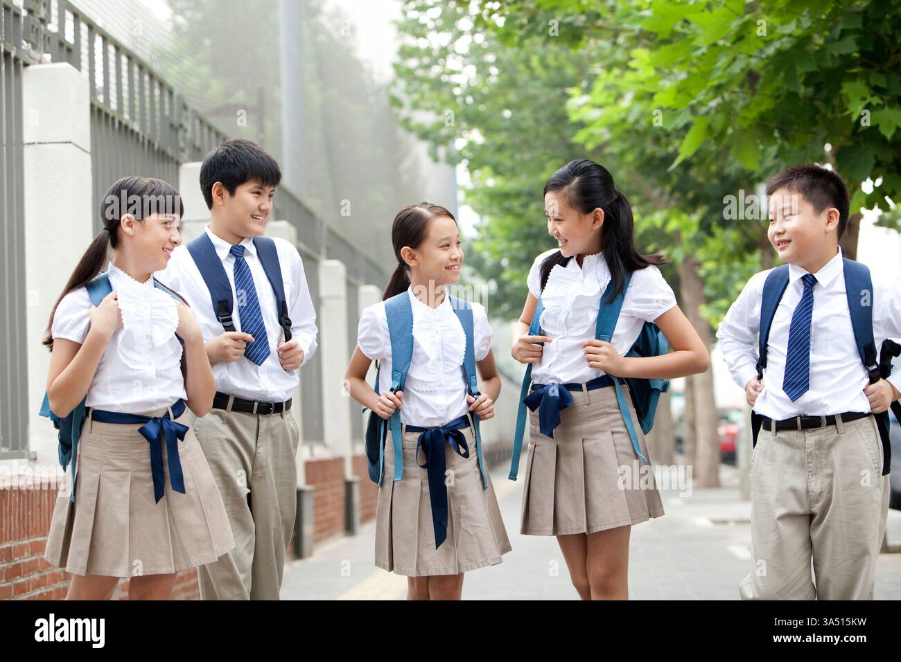 Gli studenti delle elementari cinesi sorridenti e multirazziali in uniforme camminano insieme in una strada cittadina, zaini pronti per la scuola, durante il giorno. La scena trasmette amicizia, routine quotidiane e confidenza dei primi studenti in un ambiente urbano diversificato. Da utilizzare per la copertura del ritorno alla scuola, dell'educazione multiculturale e del benessere dei giovani. Foto Stock