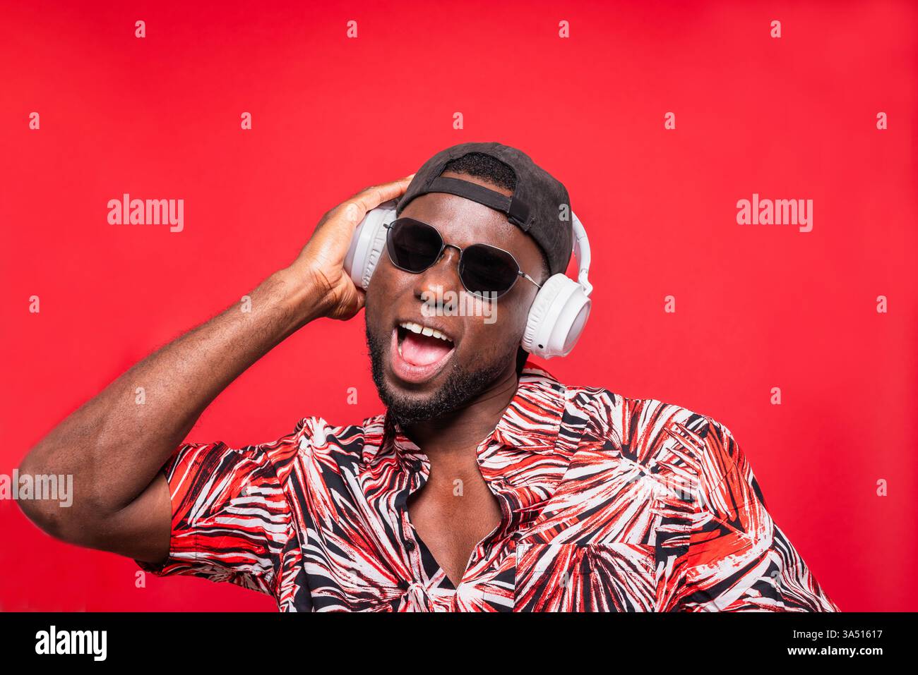 Uomo nero che indossa occhiali da sole che tocca le cuffie mentre canta in piedi su sfondo rosso Foto Stock