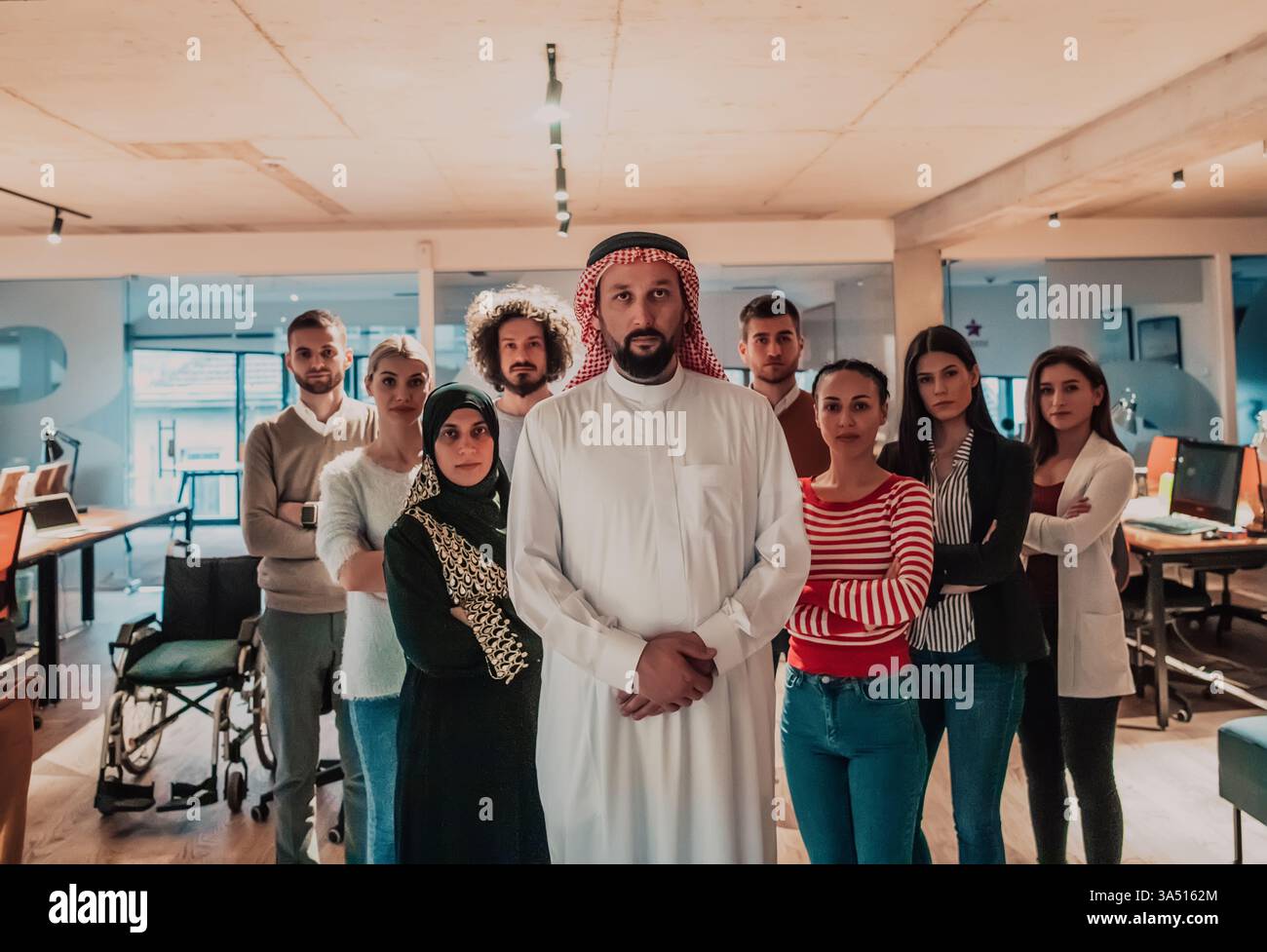 Serio uomo d'affari mediorientale che indossa kandura con le mani agganciato in piedi con diversi colleghi con le braccia incrociate contro le scrivanie in carica Foto Stock
